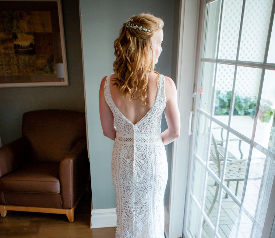 A bride-to-be looks out the window of a dressing room while showing off the back of her dress